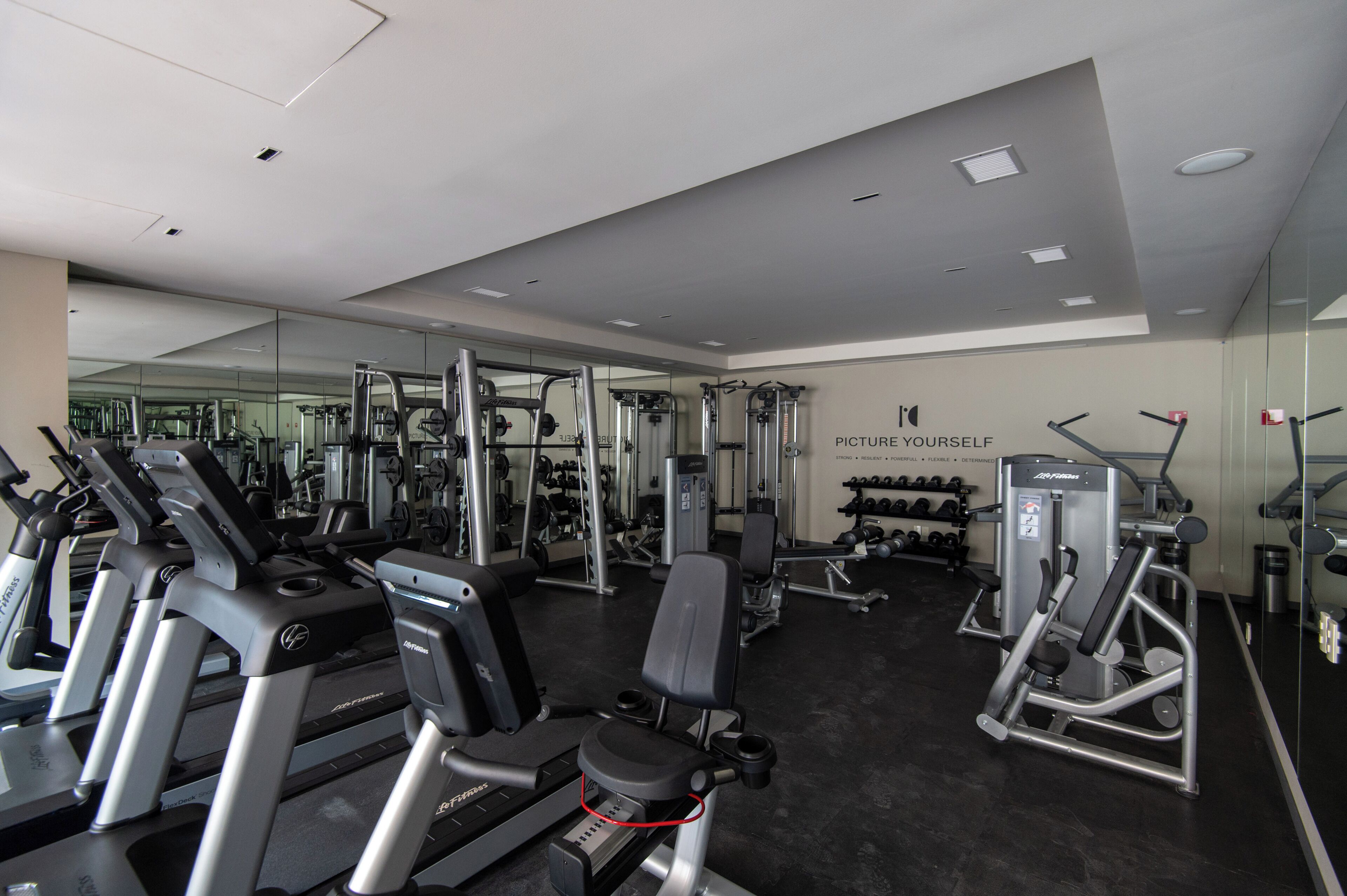Une salle de sport moderne équipée de divers appareils d’exercice, notamment des tapis de course et des appareils de musculation, ainsi qu’un grand miroir fixé au mur.
