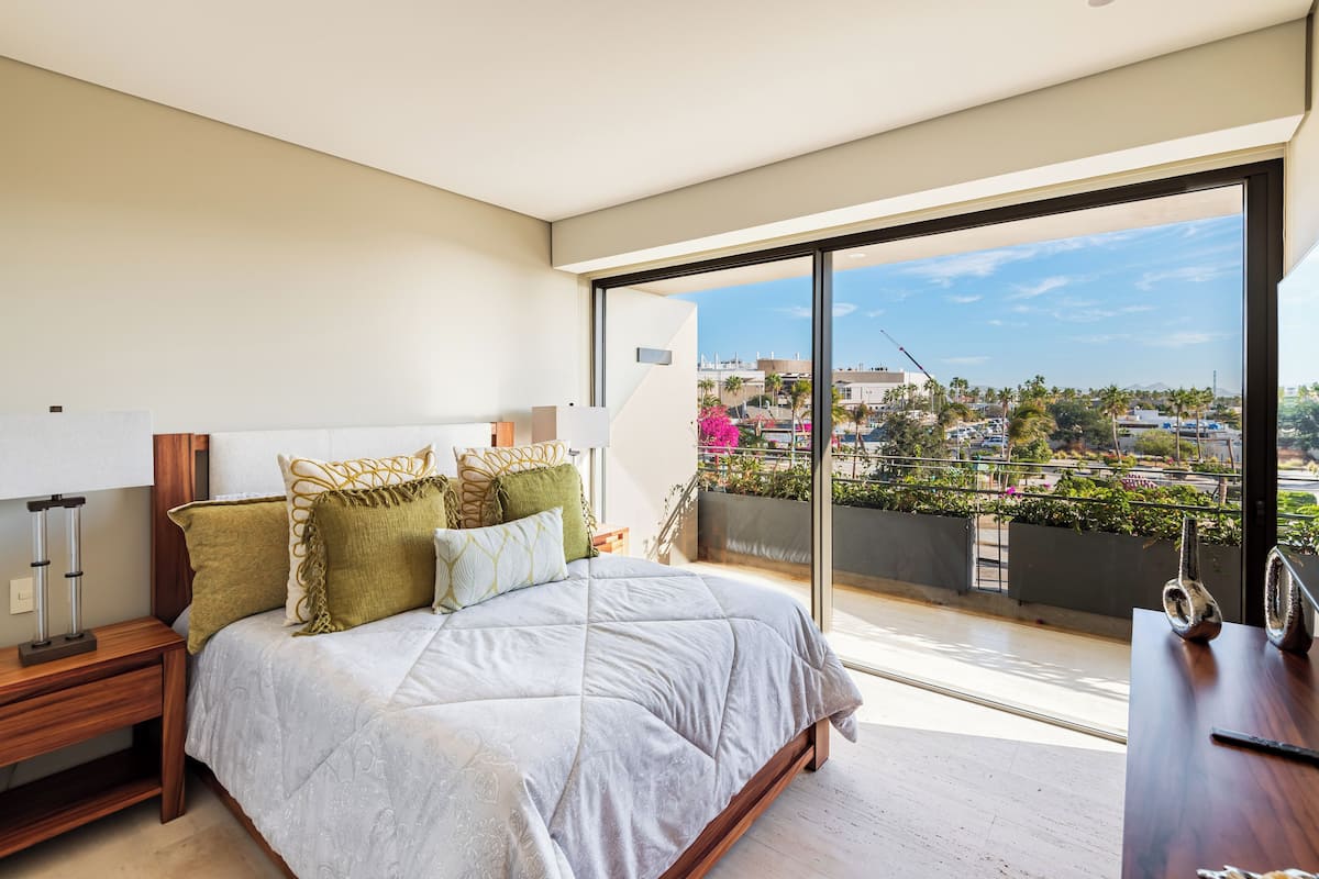 A modern bedroom with a large bed, a nightstand, and a view of a cityscape through a balcony.