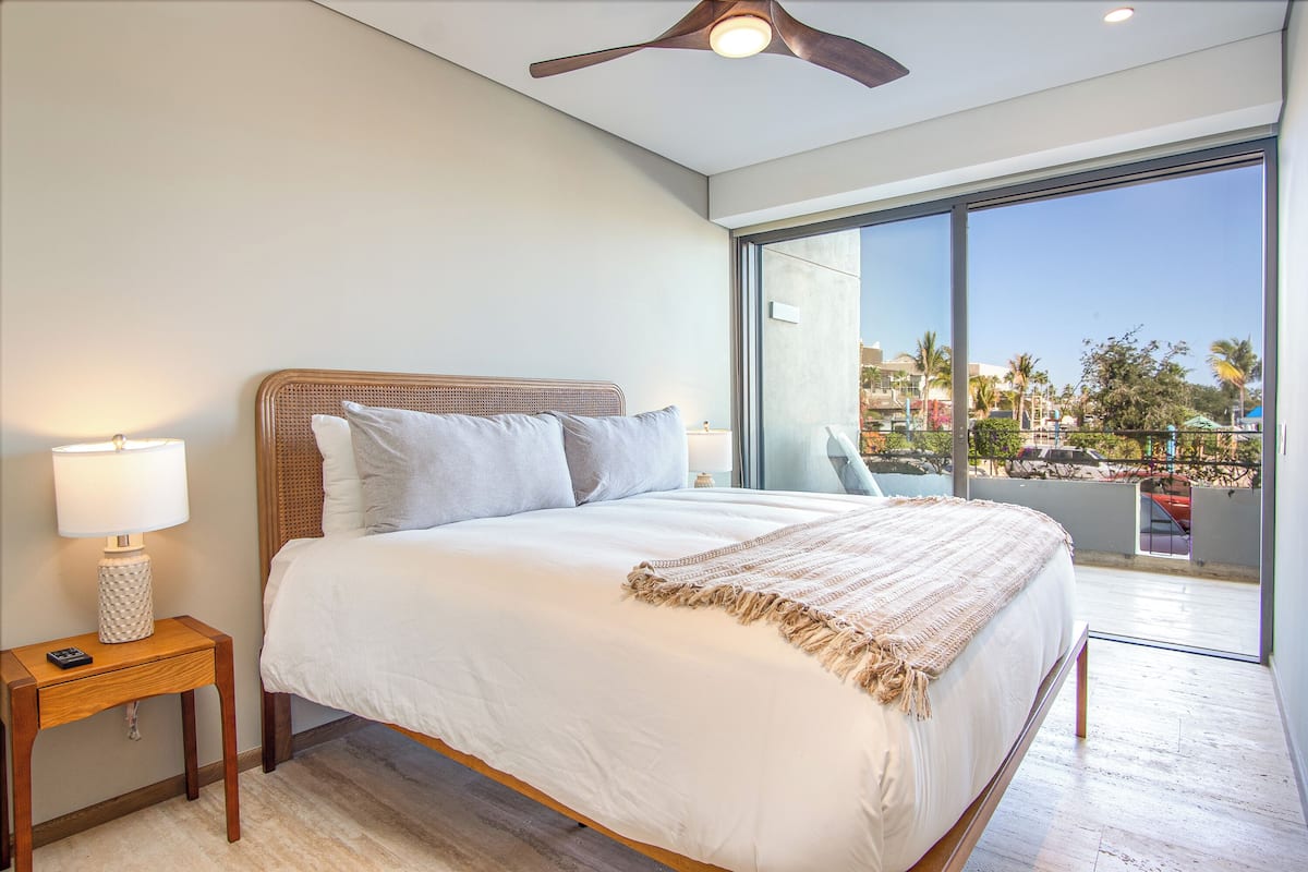 A modern hotel room with a large bed, a nightstand with a lamp, and a view of a resort area through large windows.
