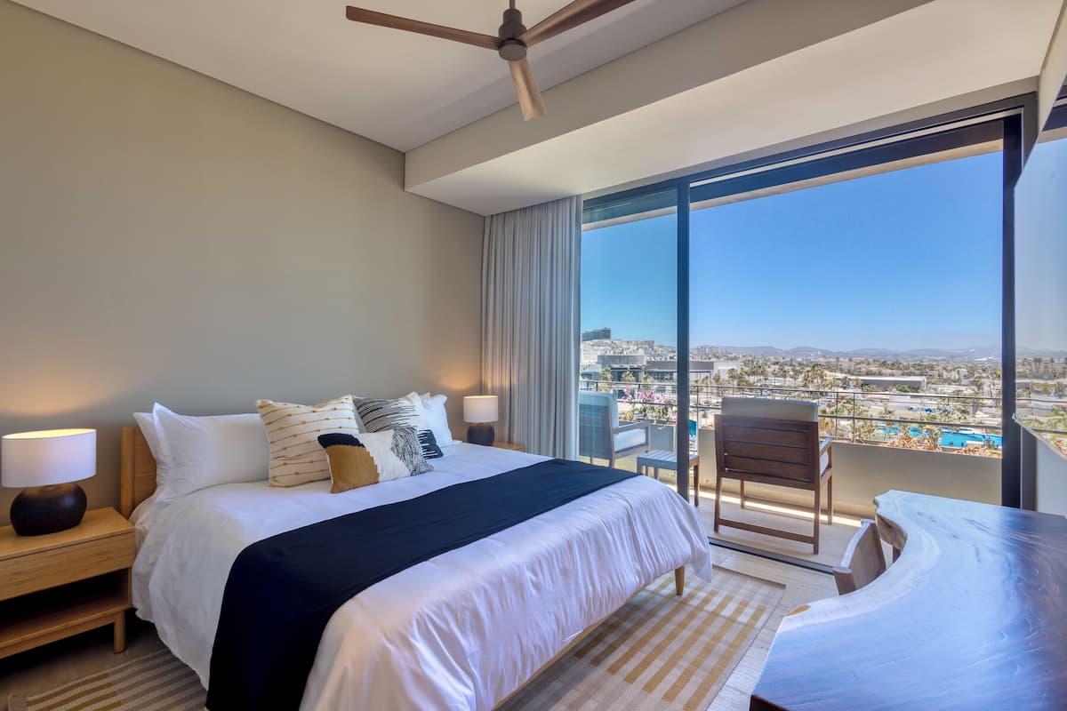 A modern hotel room with a large bed, a ceiling fan, a balcony with a chair, and a view of the city.