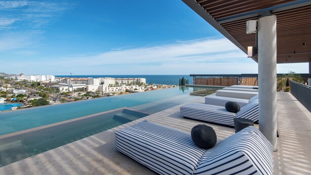 Une piscine à débordement située sur le toit, avec des chaises longues à rayures et une vue sur l’océan et le paysage urbain.