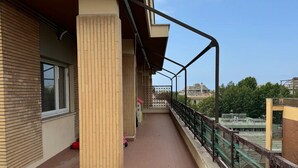 Ein überdachter Außenbereich mit gefliestem Boden, einem Metallgeländer und Blick auf Gebäude und Bäume im Hintergrund.