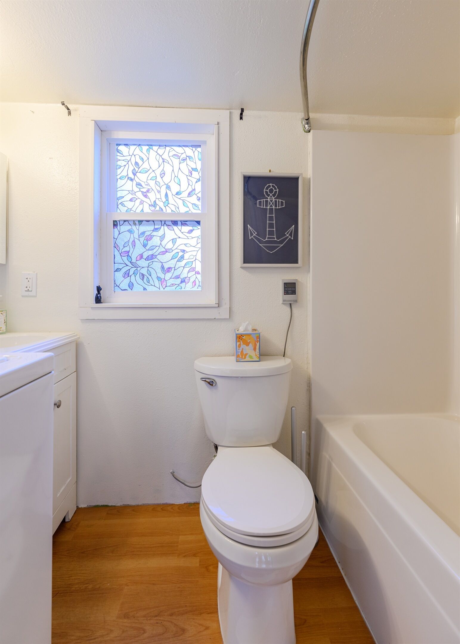 A small bathroom with a white toilet, a window with stained glass, a framed anchor artwork, and a bathtub.
