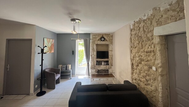 A living room with a stone wall, a black sofa, a TV, and a ceiling light. - Gîte Lavande*Clim*Terrace*Community pool (Saint-Paul-Trois-Châteaux)