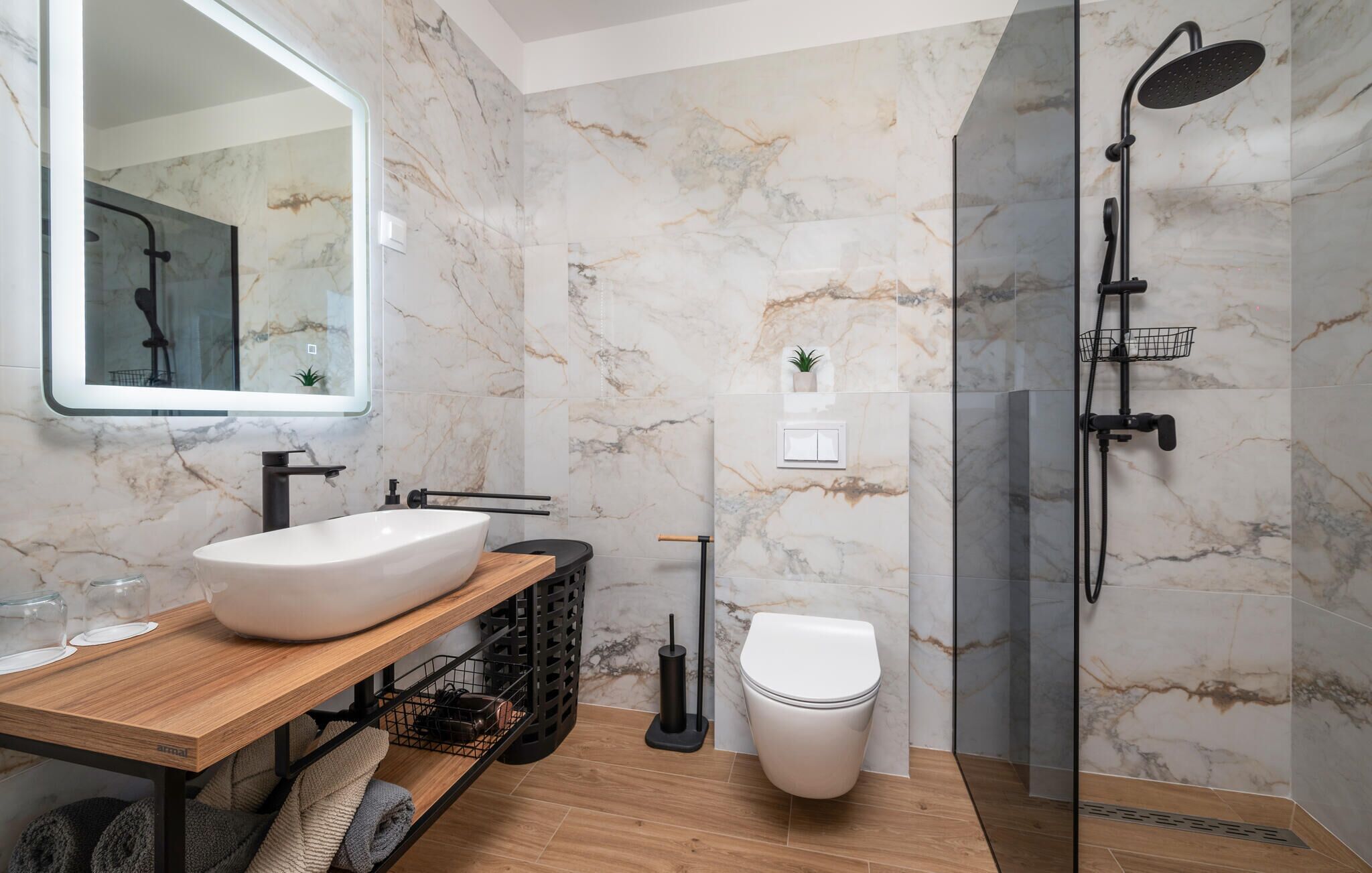 A modern bathroom with a marble wall, a wooden vanity, a white sink, a toilet, and a glass-enclosed shower area.