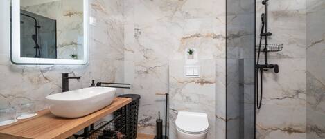 A modern bathroom with a marble wall, a wooden vanity, a white sink, a toilet, and a glass-enclosed shower area.