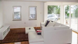 A modern living room with a white sofa, a small coffee table, and a brown rug. There are two windows with blinds, and a view of a car and a dog outside. - 2 bedroom beautiful apartment in Novi Vinodolski (Novi Vinodolski)