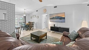 A living room with a brown leather sofa, a coffee table, a TV mounted on the wall, and a ceiling fan. - BR Tropical Oasis in Port St Lucie (Port Saint Lucie)