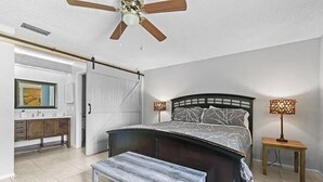 A bedroom with a curved bed, a ceiling fan, a bench, and a nightstand with a lamp. - BR Tropical Oasis in Port St Lucie (Port Saint Lucie)