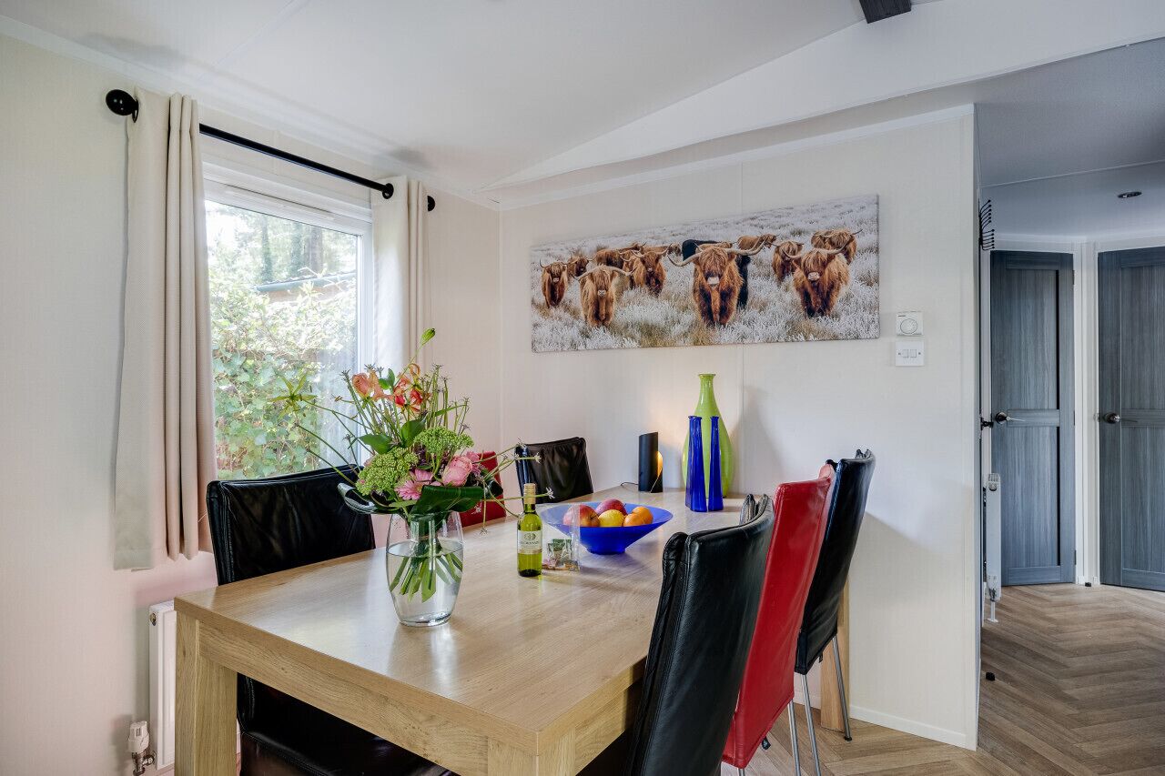 A dining area with a wooden table, chairs, a vase with flowers, a bowl of fruit, and a painting of cattle on the wall.