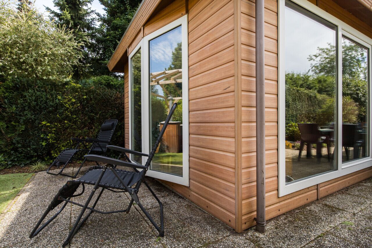A wooden cabin with large sliding glass doors, a patio with two black lounge chairs, and a visible garden area.