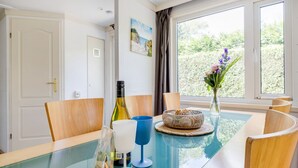 A dining area with a glass table, wooden chairs, a bottle, a vase with flowers, and a painting on the wall. - Stay With Private Hottub / Sauna (Voorthuizen)