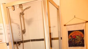 Ducha con cerramiento de vidrio, un cabezal de ducha manual, un escalón pequeño con piso perforado y un taburete blanco.