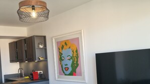 A modern living room with a round gold table, a black ottoman, a kitchen area with dark wooden cabinets, a framed pop art print, and a large flat-screen TV. - Ponant-Archipel (Valras-Plage)