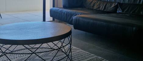 A modern living room with a black leather sofa, a round coffee table, and a patterned rug. There is a large window providing a view of the outdoors.