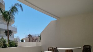 A sunlit outdoor seating area with a table and chairs, a small bowl on the table, and a view of a building and palm tree in the background. - Archipelago Garden Side (Valras-Plage)