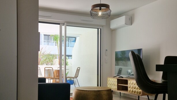 A modern living room with a large TV, a dining area with a bar stool, and a seating area with a round pouf. - Archipelago Garden Side (Valras-Plage)