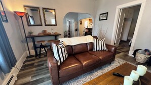 Ein Wohnzimmer mit braunem Ledersofa, zwei gestreiften Kissen, einem Couchtisch mit einem Gesellschaftsspiel und einem Essbereich mit Tisch und Stühlen.