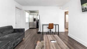 Un salon moderne avec un canapé gris foncé, une table basse en bois et un fauteuil de bureau noir. On aperçoit, au fond, une cuisine équipée d’appareils électroménagers en acier inoxydable.
