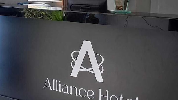 A reception desk at the Alliance Hotel with a logo, a vase of flowers, and a window in the background. - ALLIANCE HOTEL (Bauru)