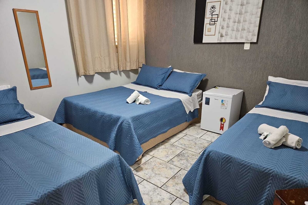Habitación de hotel con tres camas individuales, cada una con ropa de cama azul y almohadas blancas. En un lateral de la habitación hay un refrigerador blanco pequeño.