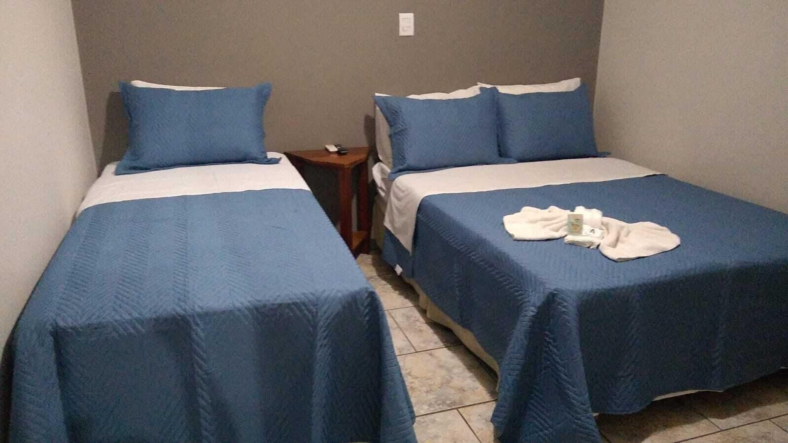 Quarto pequeno e compacto com duas camas de solteiro, cada uma com colcha e almofadas azuis. Uma pequena mesa de cabeceira de madeira fica entre as camas, com um controle remoto em cima.