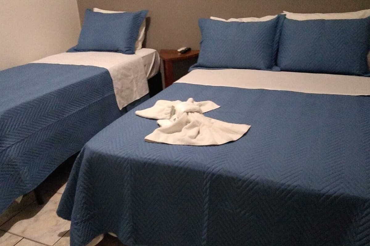 Dos camas individuales con ropa de cama azul y almohadas blancas, una mesita de noche con control remoto y una toalla blanca sobre la cama.
