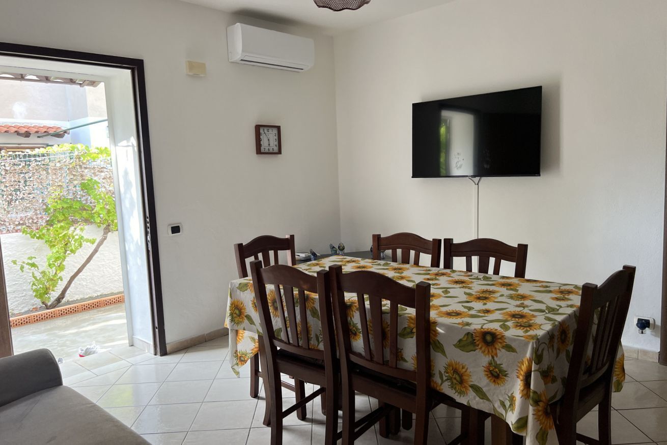 Zona pranzo con tavolo ricoperto da una tovaglia a motivi di girasoli, circondato da sedie in legno. Un televisore a schermo piatto è montato sulla parete sopra il tavolo.