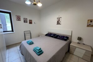 A bedroom with a bed, a nightstand, a wall-mounted lamp, and a window with blinds.
