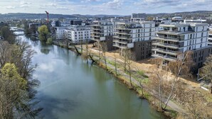 Et moderne lejlighedskompleks med flere altaner, en flod i forgrunden og en kran i baggrunden.