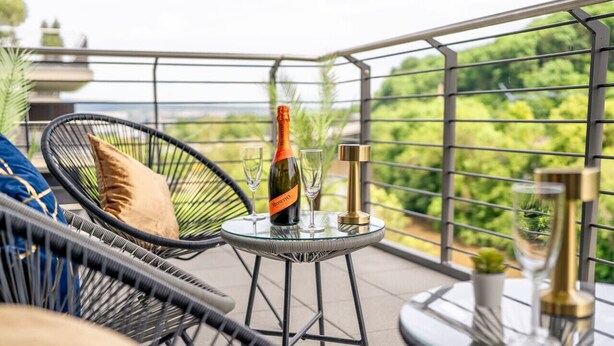 En balkon med et glasbord, to stole, en champagneflaske og en lille potteplante.