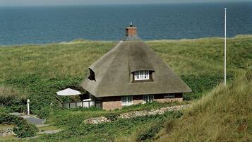 Une maison à toit de chaume, avec des fondations en pierre, située sur une colline verdoyante qui surplombe la mer.