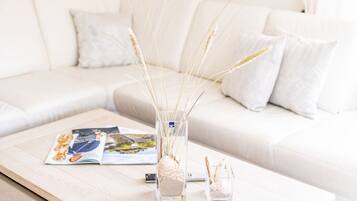 Un canapé blanc avec des coussins, une table basse en bois avec des magazines, un vase transparent contenant des plantes séchées et un petit objet décoratif.