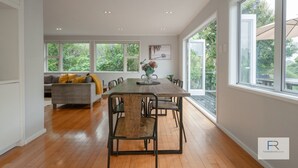 Ein geräumiger Essbereich mit Holztisch und Stühlen, einem grauen Sofa mit gelben Kissen und großen Fenstern mit Blick auf Grünflächen.