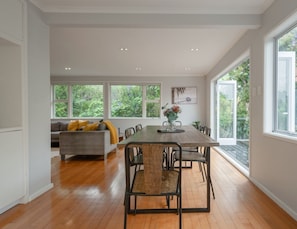 A spacious dining area with a wooden table and chairs, a grey sofa with yellow cushions, and large windows offering a view of greenery.