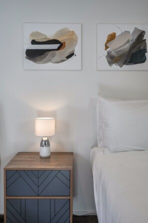 A neatly made bed with white linens, a bedside table with a lamp, and abstract artwork on the wall.