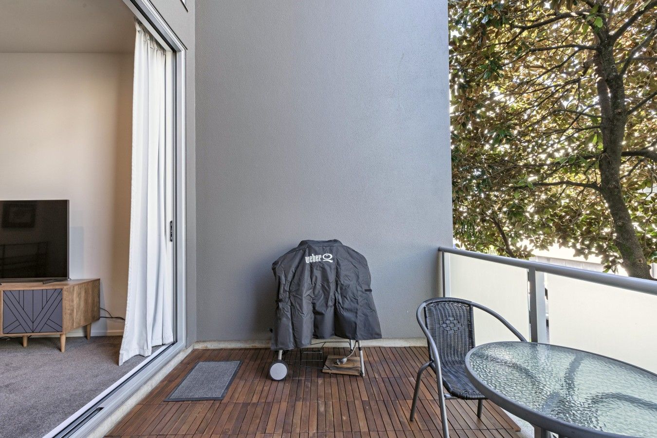 A balcony with a wooden floor, a glass table, a chair, a coat rack with a jacket, and a television on a cabinet.