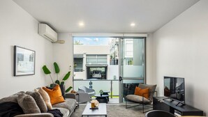 Una sala moderna con sofá, mesa de centro y televisor. Cuenta con un balcón y una puerta de vidrio que da a un espacio exterior.