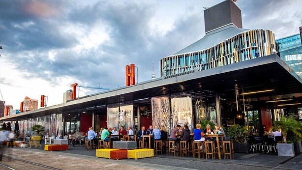 Un área de comedor al aire libre con mesas y sillas, un edificio moderno con techo curvo y un horizonte urbano de fondo.