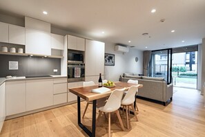 A modern kitchen with built-in appliances, a dining area with a wooden table and white chairs, and a living area with a sofa and a view of the outdoors.