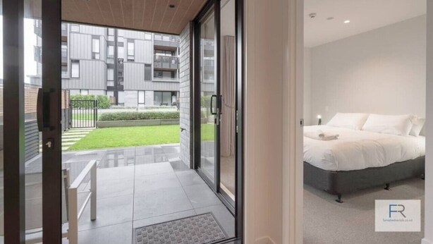 Een moderne slaapkamer met een groot bed, een glazen deur naar een balkon en uitzicht op een woongebouw.