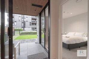 A modern bedroom with a large bed, a glass door leading to a balcony, and a view of a residential building.