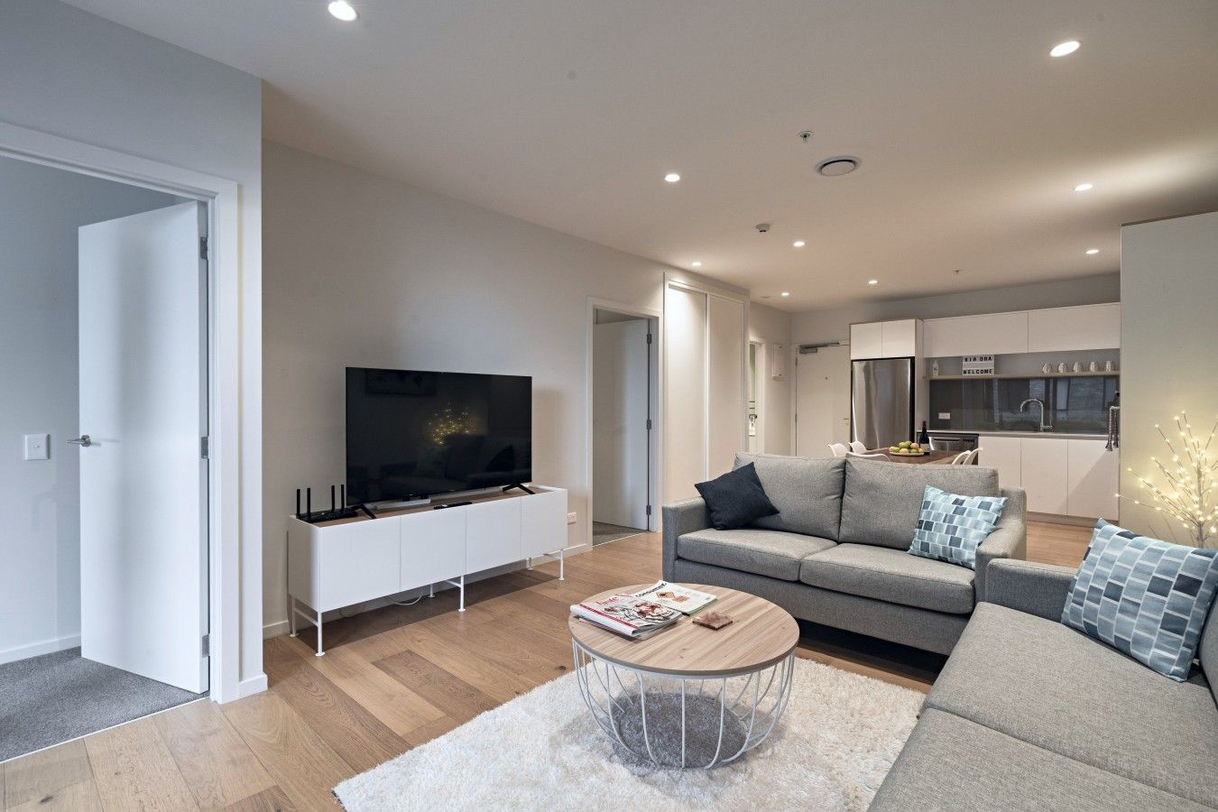 A modern living room with a flat-screen TV, a grey sofa, a coffee table, and a kitchen in the background.