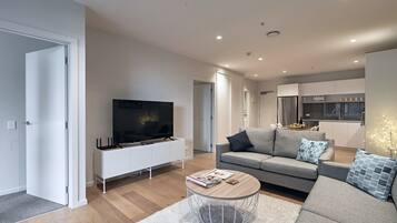 A modern living room with a flat-screen TV, a grey sofa, a coffee table, and a kitchen in the background.