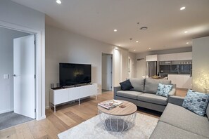 A modern living room with a flat-screen TV, a grey sofa, a coffee table, and a kitchen in the background.
