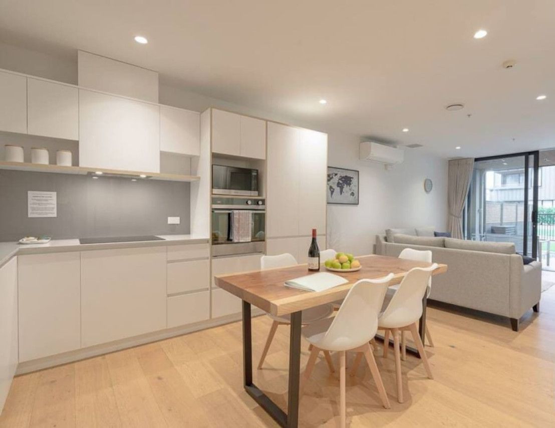 A modern kitchen with white cabinets, a wooden dining table, and a sofa in the adjacent living area.