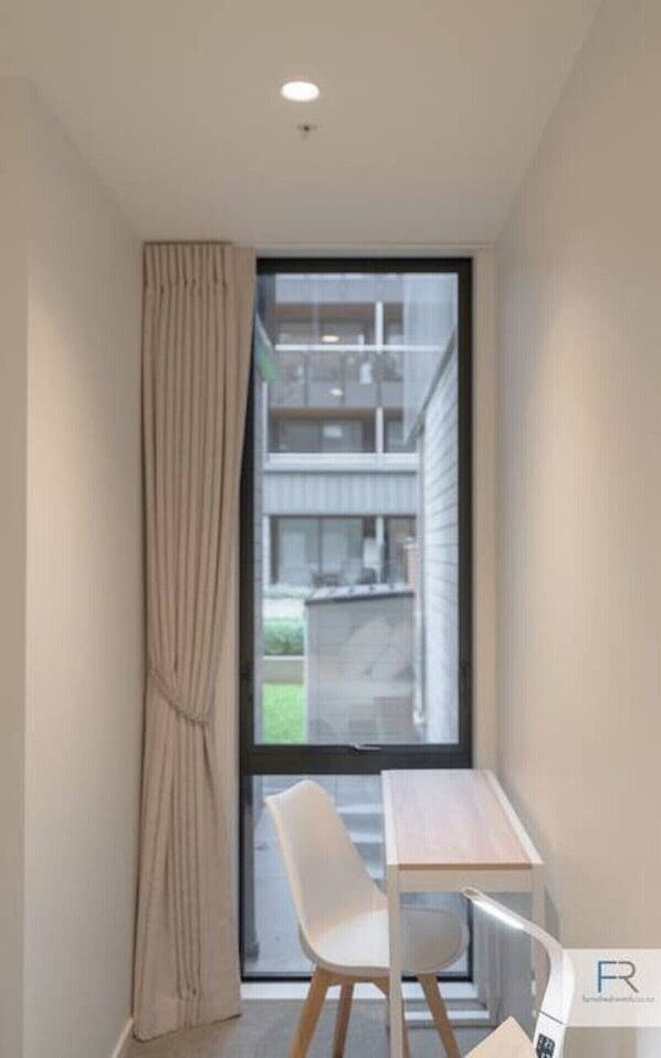 A small, modern room with a single table and chair, a large window with a view of a building, and a curtain on the left.