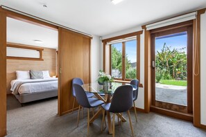 A room with a bed, a dining table with chairs, and a large window with a view of greenery.