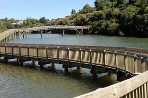 A wooden bridge over water with a railing, surrounded by trees and a clear sky. - Comfortable Cosy Apartment| by Furnished Rentals (Auckland)