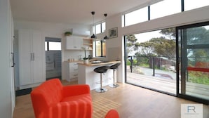 A modern kitchen with white cabinets, a wooden countertop, and a bar area with stools. There is a large sliding glass door leading to an outdoor area with a view of trees and a fence. - Cosy Tiny Home with Ocean Views|by Furnished Rentals (Auckland)